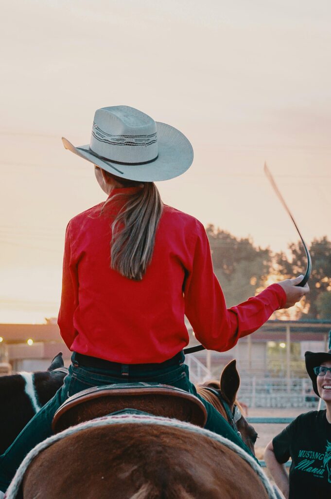 Back view of a cowgirl in a red shirt and cowboy hat riding a horse during sunset at a rodeo.